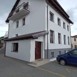 Dwukondygnacyjny budynek z balkonem i brązowymi ramami okiennymi, świeżo pomalowany na biało-szaro, z brukowanym podjazdem i zaparkowanym niebieskim samochodem.