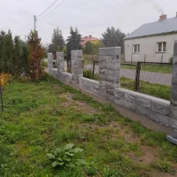 Nowo wybudowane ogrodzenie z szarych, betonowych bloczków o fakturze kamienia, z widocznymi słupkami, oddzielające posesję z zielonym trawnikiem od sąsiedniej nieruchomości z widocznym domem...