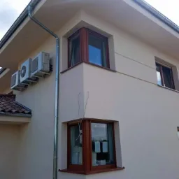 Jasna elewacja domu jednorodzinnego z brązowymi oknami, widoczne jednostki klimatyzacyjne i rynna spustowa, częściowo widoczny dach z czerwoną dachówką.