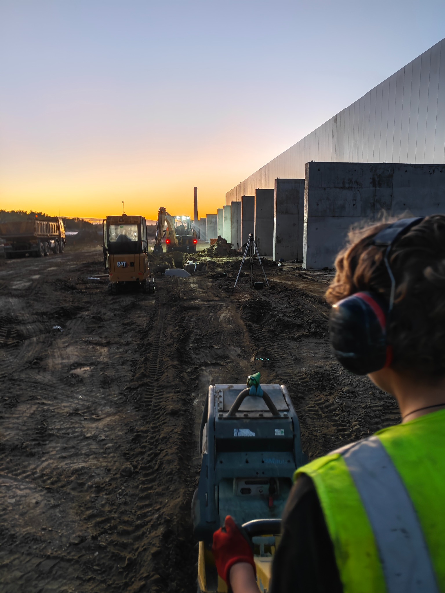 Pracownik w kamizelce odblaskowej obsługuje zagęszczarkę gruntu na placu budowy hali usługowej o świcie, w tle widoczny miniładowarka CAT, wykop, ciężarówka i budynek w konstrukcji.