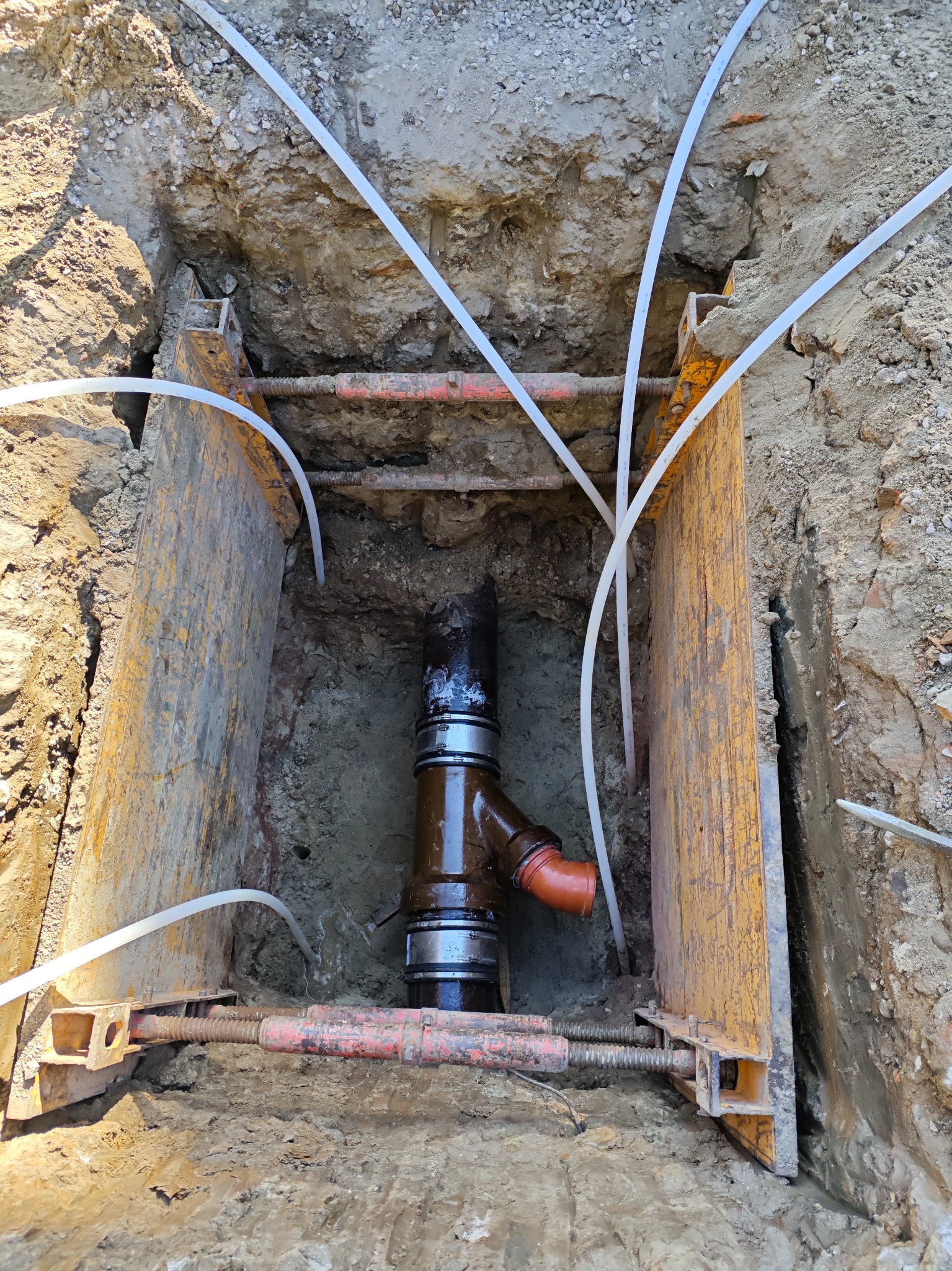 Głęboki wykop zabezpieczony żółtymi szalunkami stalowymi, wewnątrz pionowy rurociąg z brązowymi elementami i pomarańczowym kolankiem, otoczony białymi rurkami, ziemia na ścianach wykopu wilgotna...