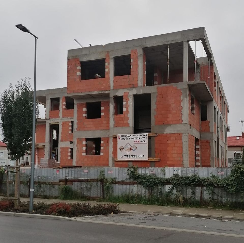 Surowy stan budynku wielorodzinnego z czerwonej cegły i betonu, widoczne otwory okienne i balkonowe, tablica reklamowa firmy budowlanej na ogrodzeniu.