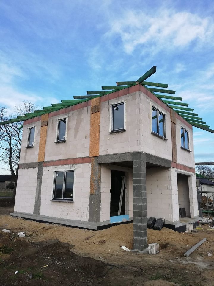 Dwukondygnacyjny dom w trakcie budowy z białych bloczków, widoczne okna z czarnymi ramami, przygotowane elementy konstrukcji dachu z zielonego drewna, na zewnątrz ziemia i piasek.