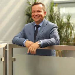 Uśmiechnięty mężczyzna w niebieskim garniturze i krawacie w czerwone wzory, oparty o szklaną balustradę, z zegarkiem na lewym nadgarstku, na tle roślin doniczkowych i żaluzji.