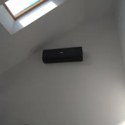 Czarny klimatyzator Haier zamontowany na skośnej ścianie pod oknem dachowym w jasnym pomieszczeniu.