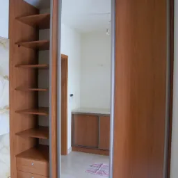 Drewniana szafa z przesuwnymi drzwiami i lustrem, z otwartymi półkami po lewej stronie, widok na wnętrze pomieszczenia z szafką i dywanikiem w odbiciu lustrzanym.