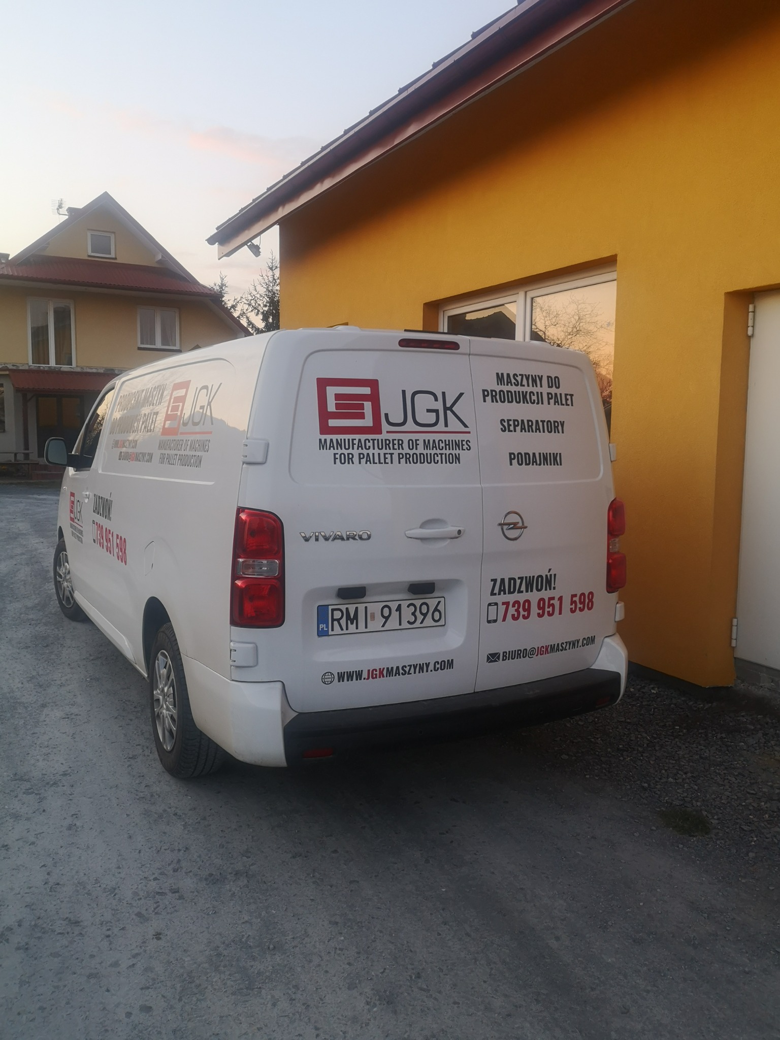 Biały samochód dostawczy firmy JGK z reklamą maszyn do produkcji palet, separatorów i podajników, zaparkowany przed żółtym budynkiem w słoneczny dzień.