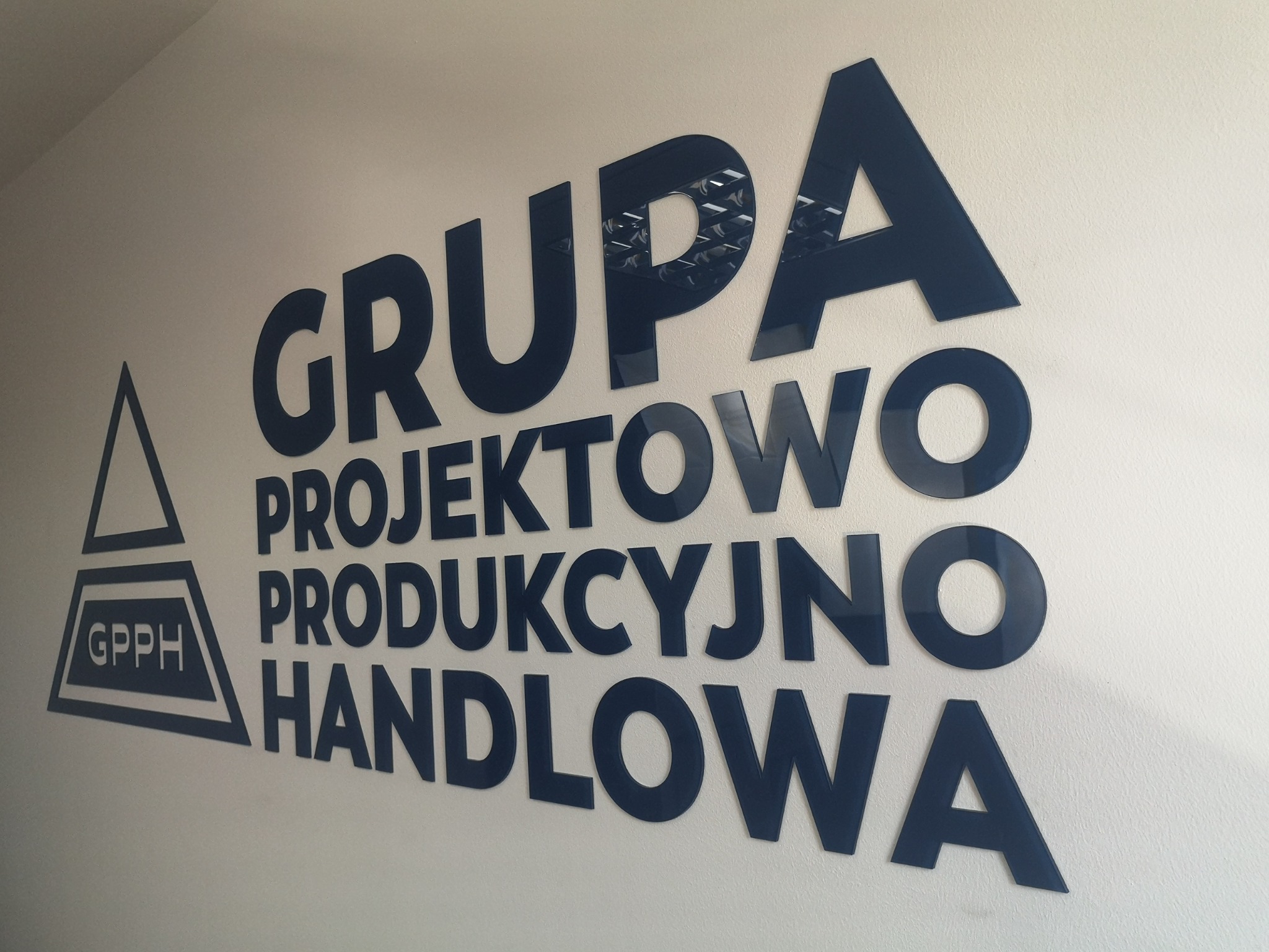 Niebieski napis 'GRUPA PROJEKTOWO PRODUKCYJNO HANDLOWA' oraz logo GPPPH umieszczone na jasnej ścianie, wykonane z błyszczącego tworzywa.