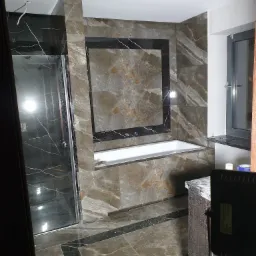 Wykończona łazienka z wanną obudowaną płytkami imitującymi marmur w odcieniach brązu i szarości, z kabiną prysznicową po lewej stronie i oknem po prawej, widoczne wystające kable z sufitu.