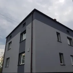 Dwukolorowa elewacja budynku mieszkalnego w odcieniach szarości, widoczna z dołu pod pochmurnym niebem, z numerem '2' na ścianie i brązową rynną.