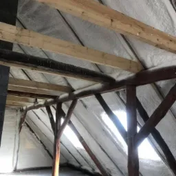 Poddasze ocieplone pianką, widoczne drewniane elementy konstrukcyjne, szczeliny okienne w skośnej ścianie, światło słoneczne wpadające do wnętrza.