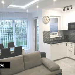 Jasne wnętrze mieszkania z aneksem kuchennym: białe meble kuchenne z czarnym blatem i mozaiką nad zlewem, zegar na ścianie, stół z krzesłami przy dużym oknie z żaluzjami pionowymi, widok z salonu...