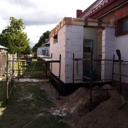 Wznoszenie ścian z bloczków betonowych na fundamencie zabezpieczonym izolacją, widoczne rusztowania i narzędzia na placu budowy przy ceglanym budynku.