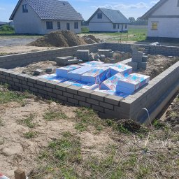 BUDAN- Usługi Ogólnobudowlane - Wzniesione mury fundamentowe z bloczków betonowych, zabezpieczone izolacją termiczną w postaci białych płyt styropianowych. W tle budynki w stanie surowym.