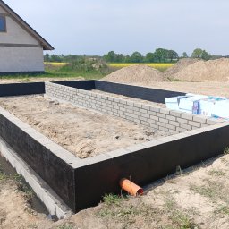 BUDAN- Usługi Ogólnobudowlane - Wykop pod fundamenty z izolacją pionową i częściowo wymurowanymi ścianami z bloczków. W tle widać budynek w stanie surowym otwartym i żółte pola.