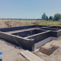 BUDAN- Usługi Ogólnobudowlane - Zabezpieczone fundamenty domu jednorodzinnego z wystającymi prętami zbrojeniowymi, widok z góry na tle krajobrazu z wiatrakami i zaparkowanymi autami.