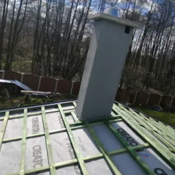 Szary komin z metalowym daszkiem wystaje z dachu w trakcie budowy, widoczne drewniane łaty i membrana dachowa marki CREATON, w tle las i ogrodzenie.