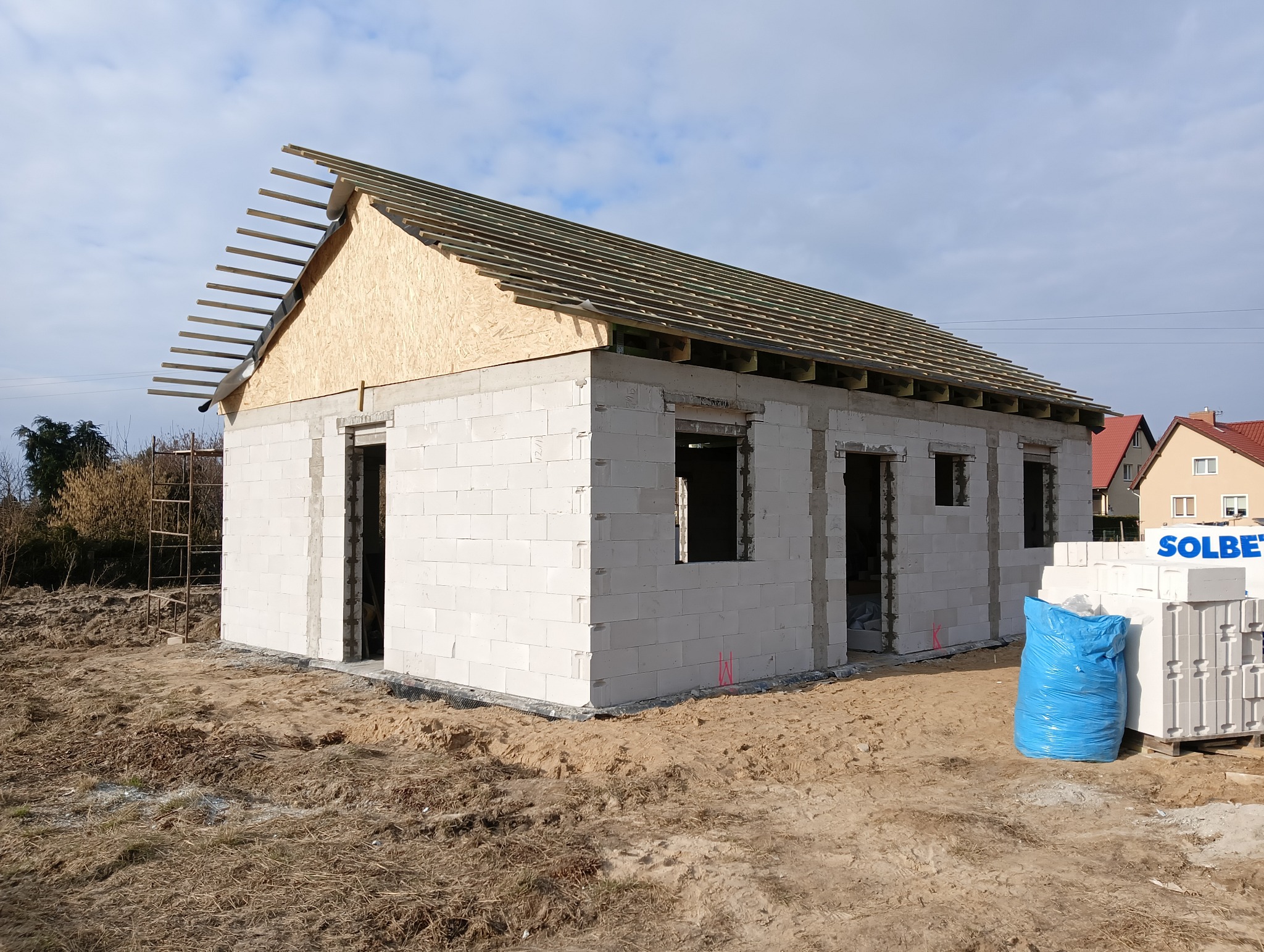 Niedokończony dom z białych bloczków, widoczne otwory okienne i drzwiowe, konstrukcja dachu z desek i łat. Na pierwszym planie paleta z bloczkami Solbet.