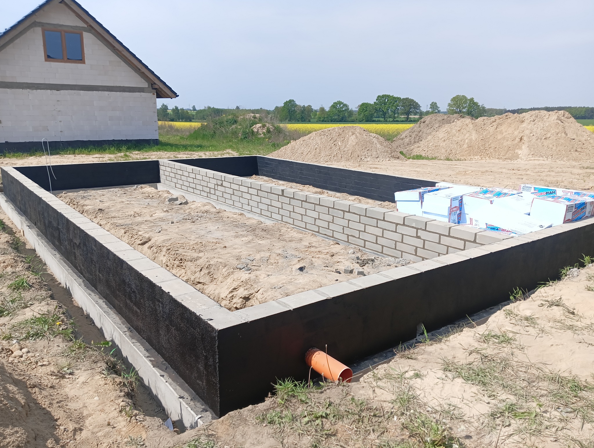 Wykop pod fundamenty z izolacją pionową i częściowo wymurowanymi ścianami z bloczków. W tle widać budynek w stanie surowym otwartym i żółte pola.