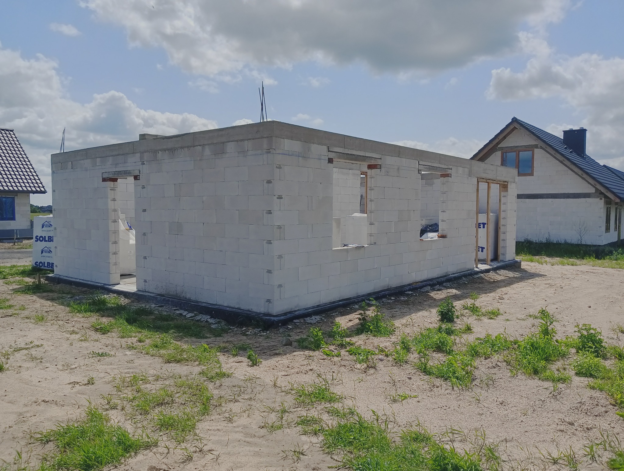 Budowa domu w stanie surowym otwartym z betonu komórkowego. Widoczne ściany, otwory okienne i drzwiowe. W tle drugi budynek z ciemną dachówką.