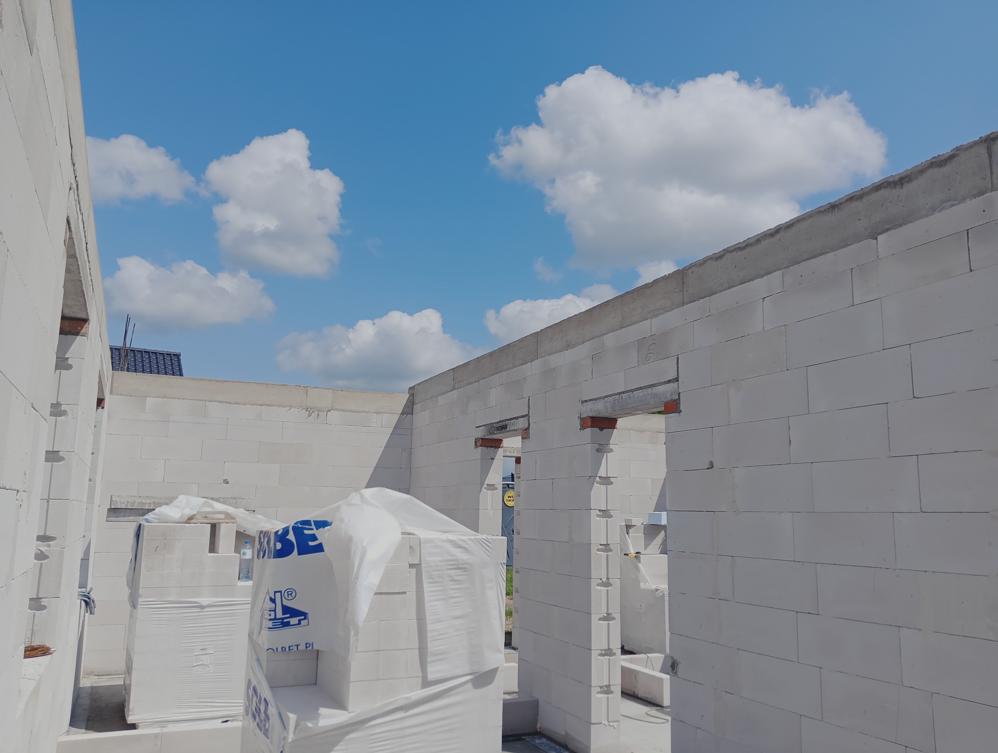 Wnętrze domu w trakcie budowy z białych bloczków, widoczne otwory okienne i drzwiowe, paleta z materiałami budowlanymi w folii ochronnej, błękitne niebo w tle.