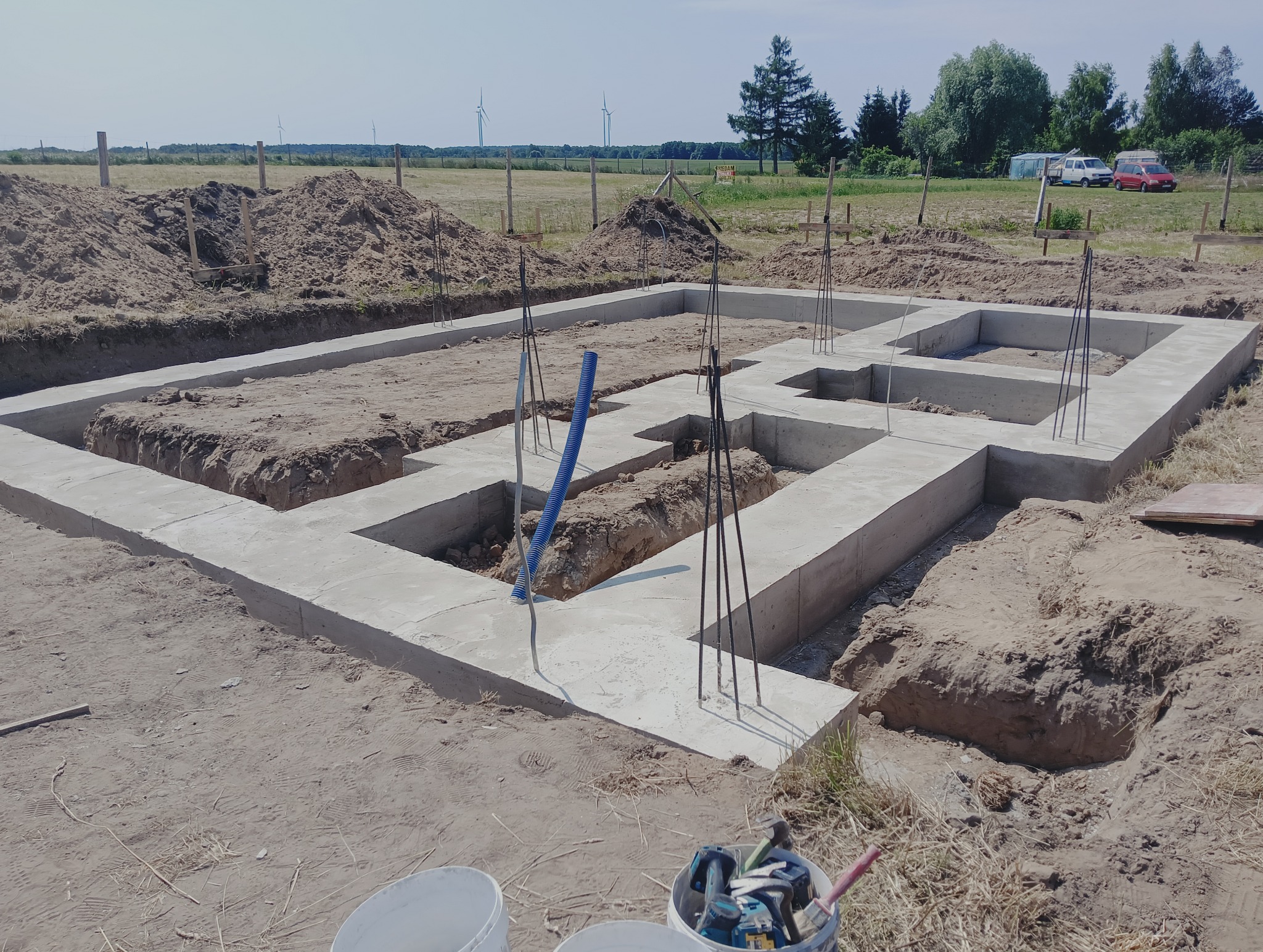 Zalane fundamenty domu jednorodzinnego z wystającymi prętami zbrojeniowymi, widoczne narzędzia w wiadrze, w tle krajobraz z turbinami wiatrowymi.