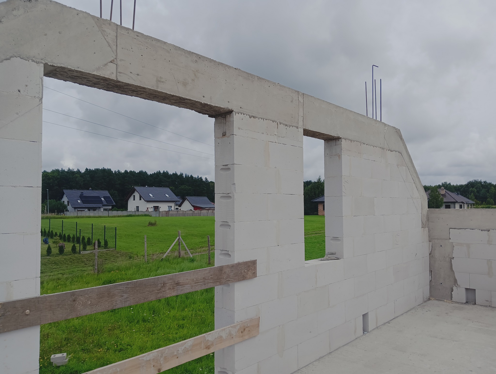 Ściana domu w trakcie budowy z betonu komórkowego, widoczne otwory okienne i zbrojenie. W tle zieleń i zabudowa jednorodzinna.