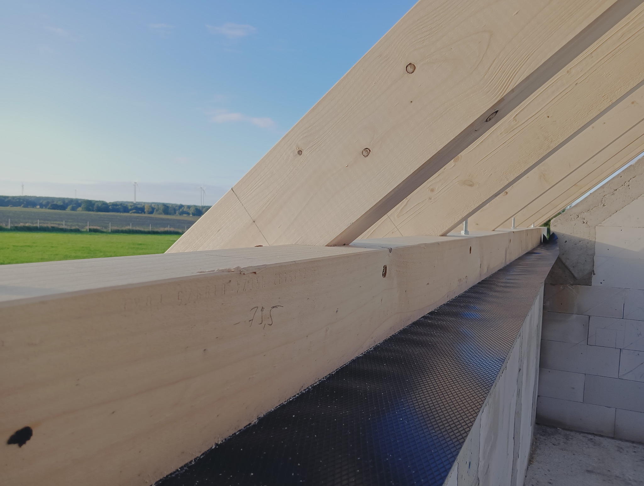 Konstrukcja drewniana dachu w trakcie budowy, widoczne elementy więźby i ściany z betonu komórkowego, na tle zielonego pola i błękitnego nieba.