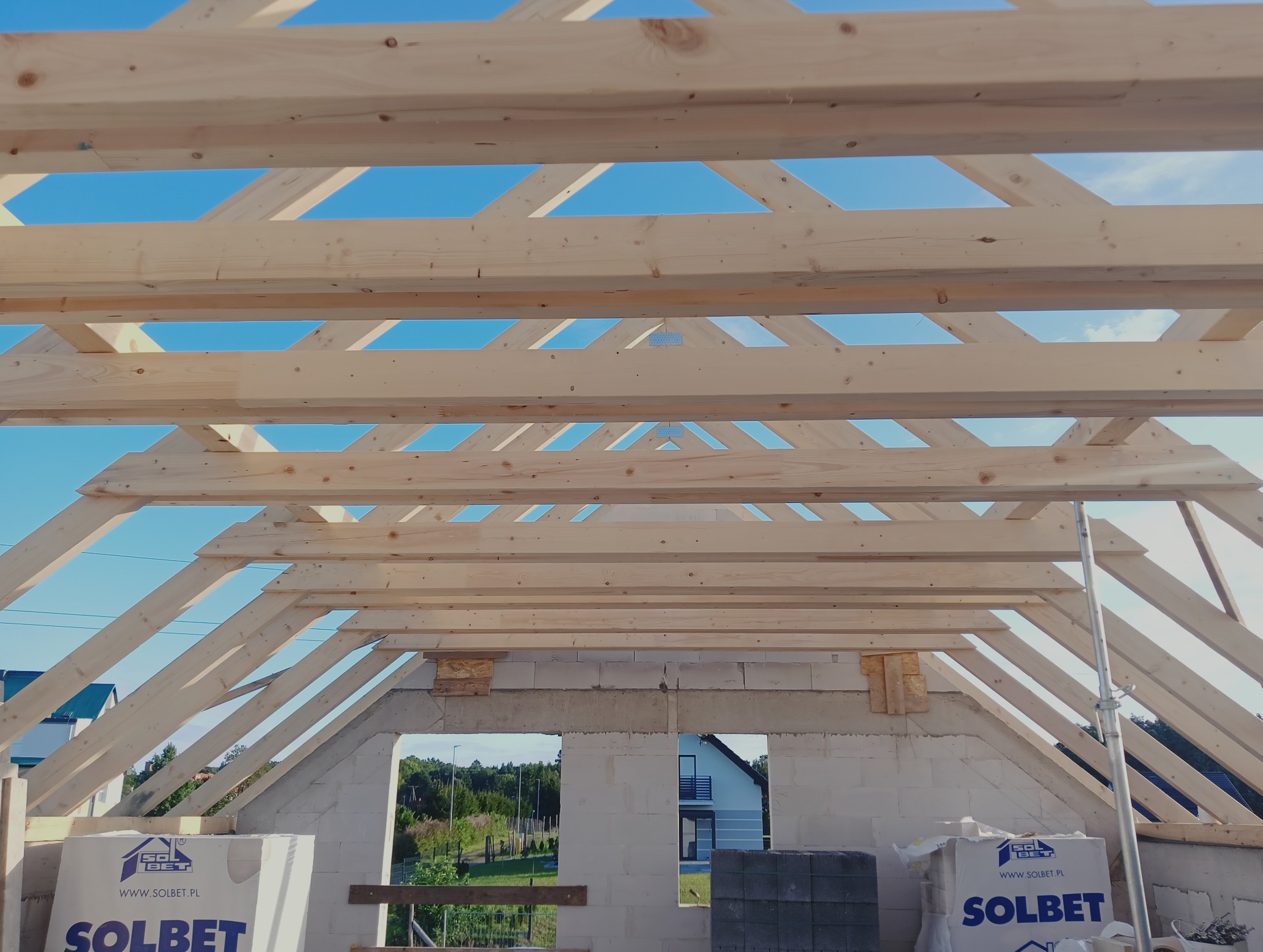 Konstrukcja drewniana dachu w trakcie budowy domu, widok z wnętrza. Białe bloczki Solbet na ścianach, błękitne niebo w tle. Widoczne elementy rusztowania.