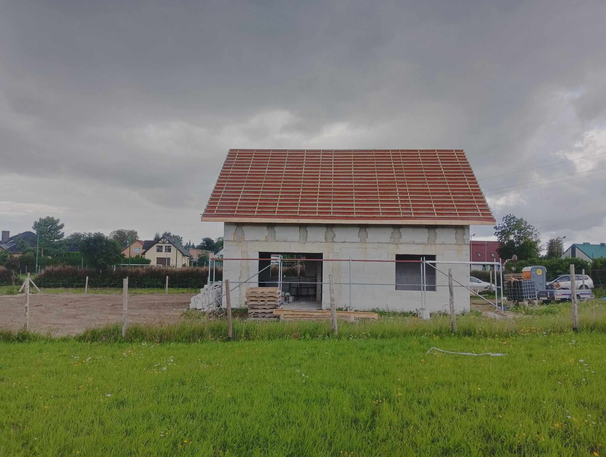 Niedokończony dom z pustaków z częściowo ułożonym czerwonym dachem na tle pochmurnego nieba i zielonej trawy. Widoczne rusztowania i materiały budowlane.