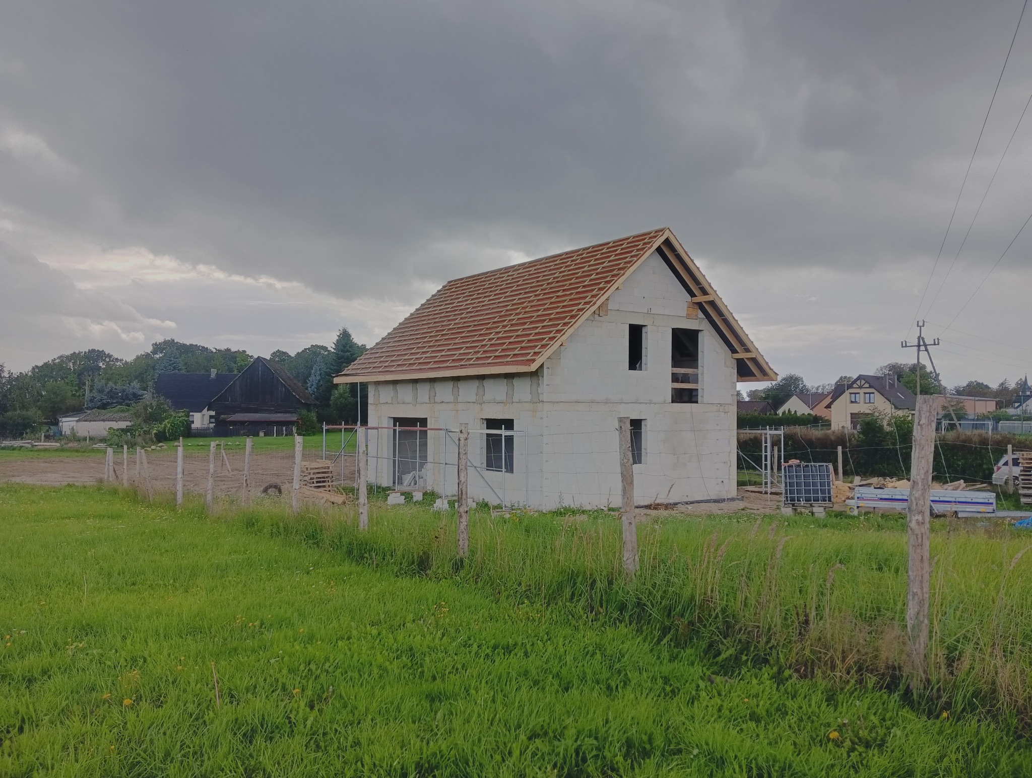 Surowy dom z pustaków z częściowo ułożoną dachówką, otoczony zielonym polem i drewnianym ogrodzeniem. Pochmurne niebo w tle. Widoczne elementy konstrukcyjne dachu.