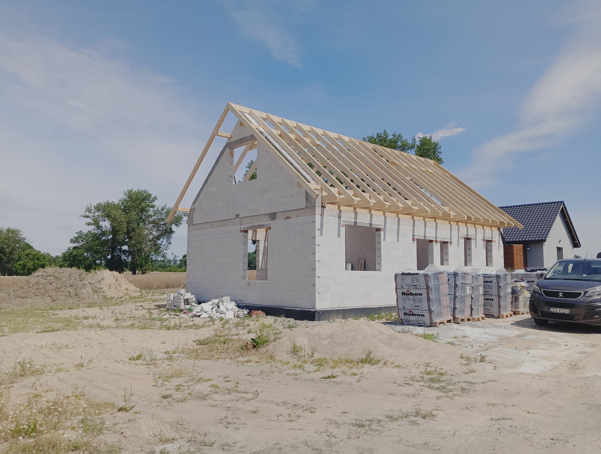 Budowa domu z białych bloczków: ściany wzniesione, więźba dachowa zamontowana, palety z bloczkami Roben obok. Widoczny samochód i sąsiedni dom w tle.