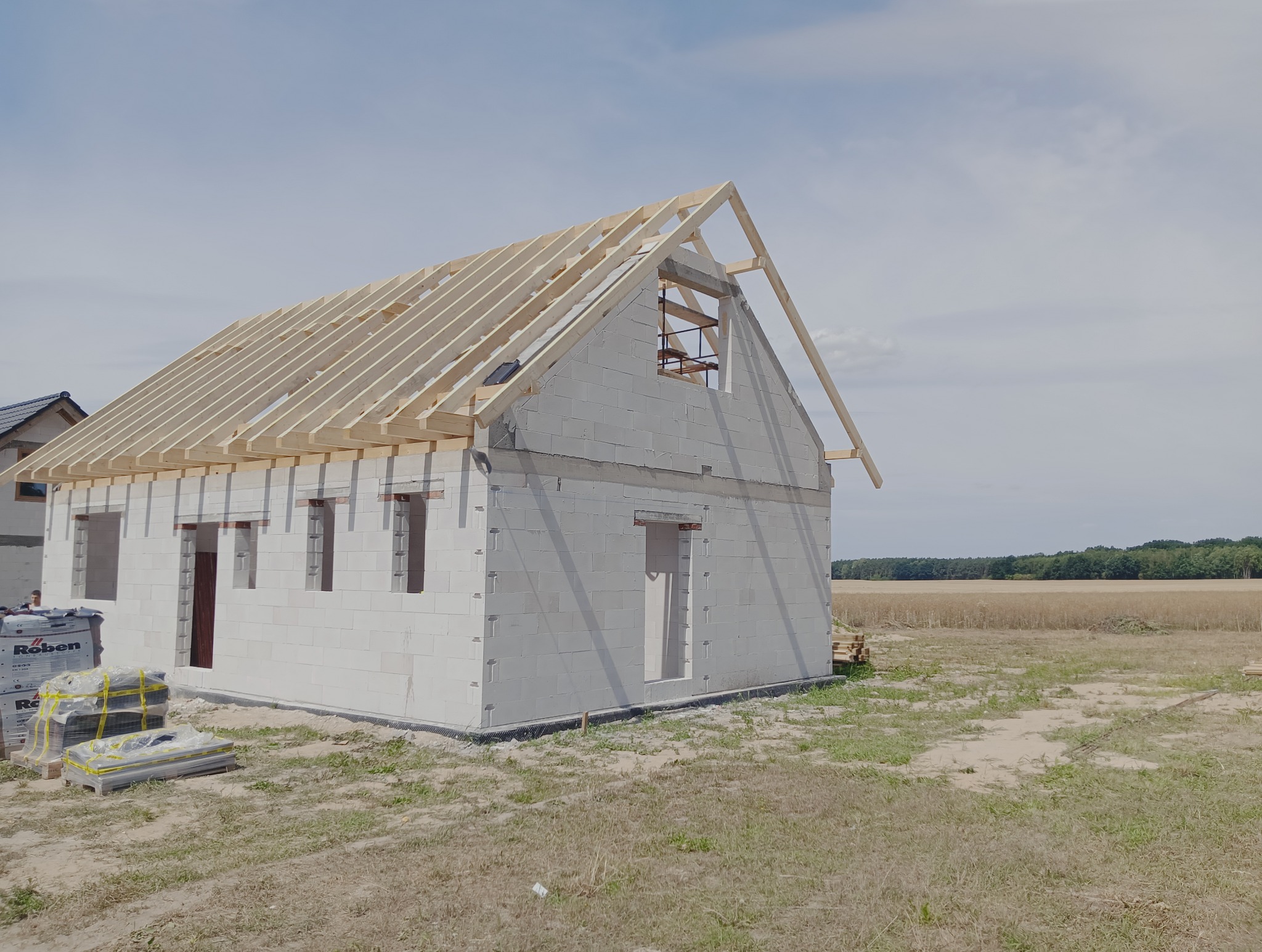 Budowa domu w stanie surowym otwartym: białe ściany z betonu komórkowego, drewniana konstrukcja dachu, pakiety bloczków Roben obok. Pola i las w tle.