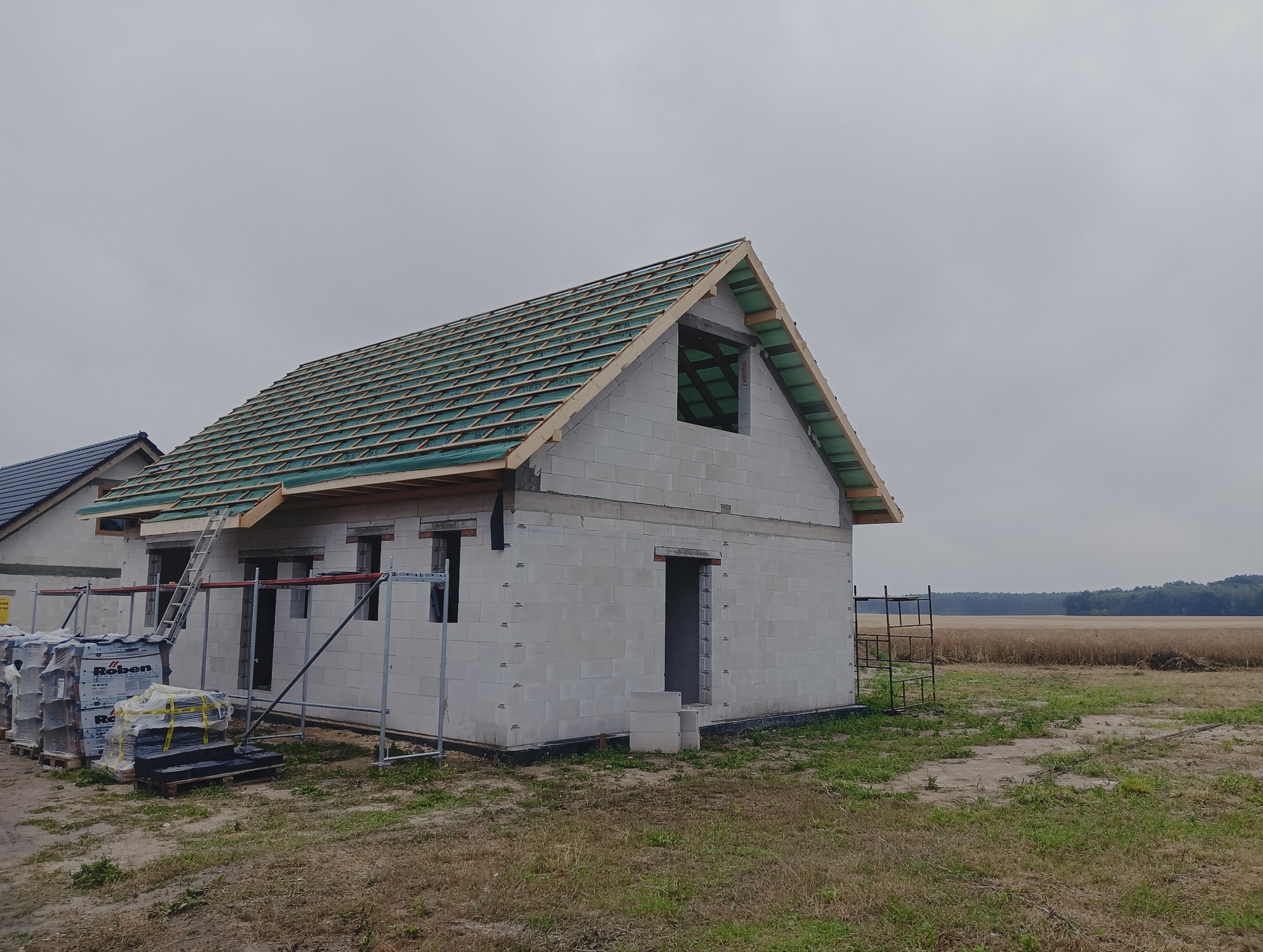 Surowy stan domu z betonu komórkowego z drewnianą konstrukcją dachu i zieloną membraną, na tle pola i lasu w pochmurny dzień. Widoczne rusztowania i materiały budowlane.