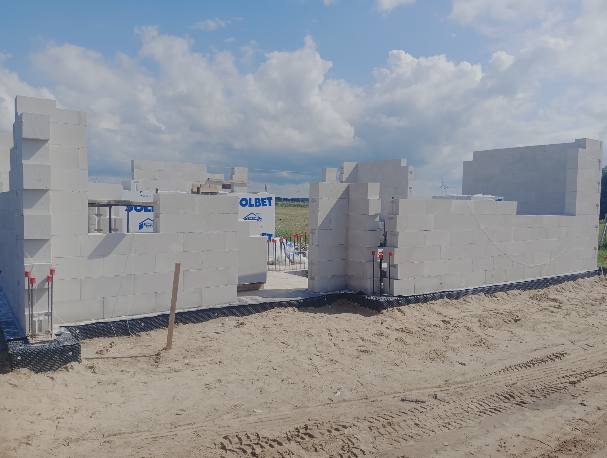 Wznoszenie ścian domu z białych bloczków, widoczna izolacja fundamentów i zbrojenia. W tle błękitne niebo z chmurami i pole z turbiną wiatrową.