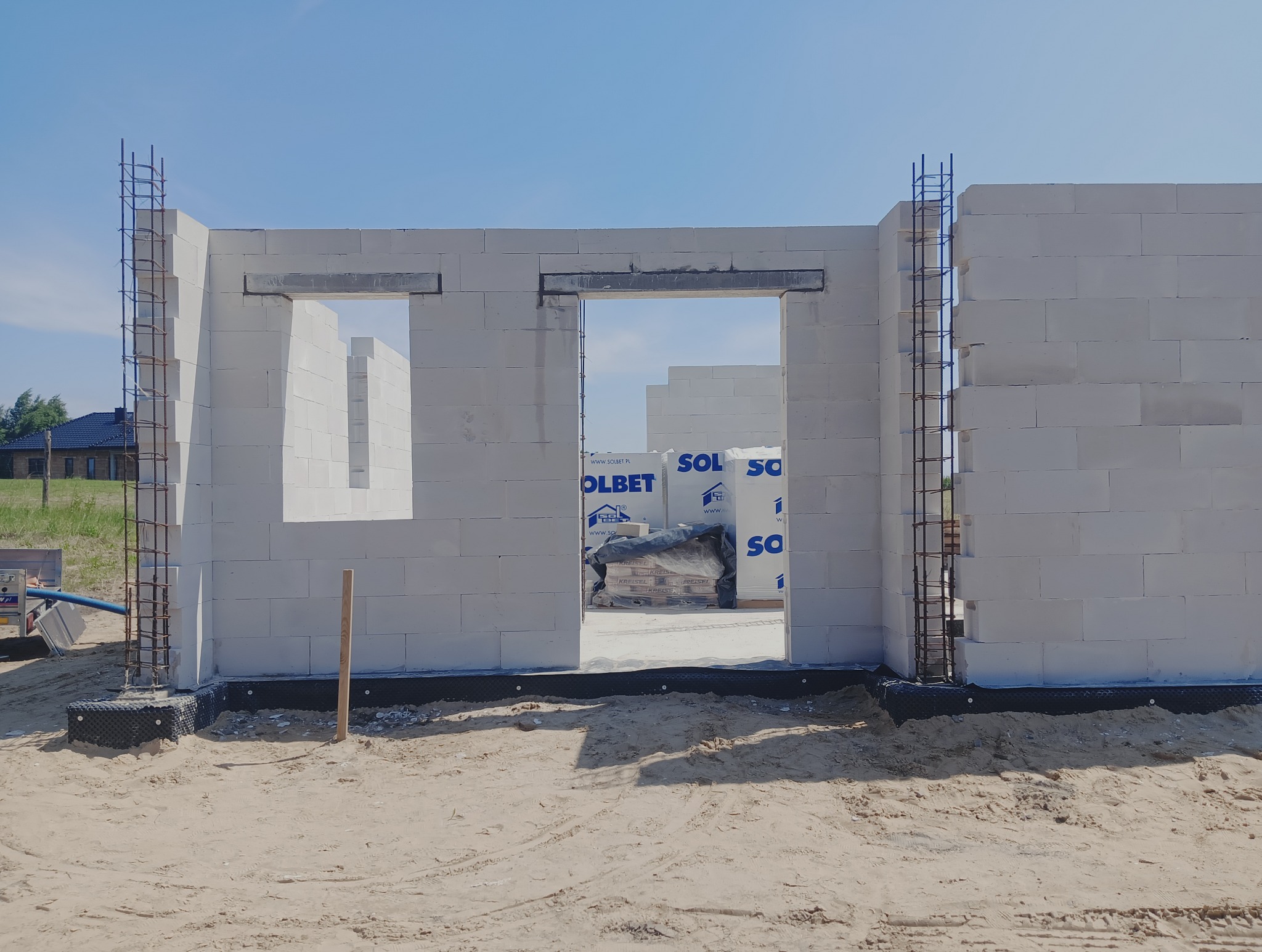 Wznoszone ściany domu z betonu komórkowego Solbet, zbrojone narożniki. Widoczne otwory okienne i drzwiowe, materiały budowlane w tle. Słoneczny dzień.