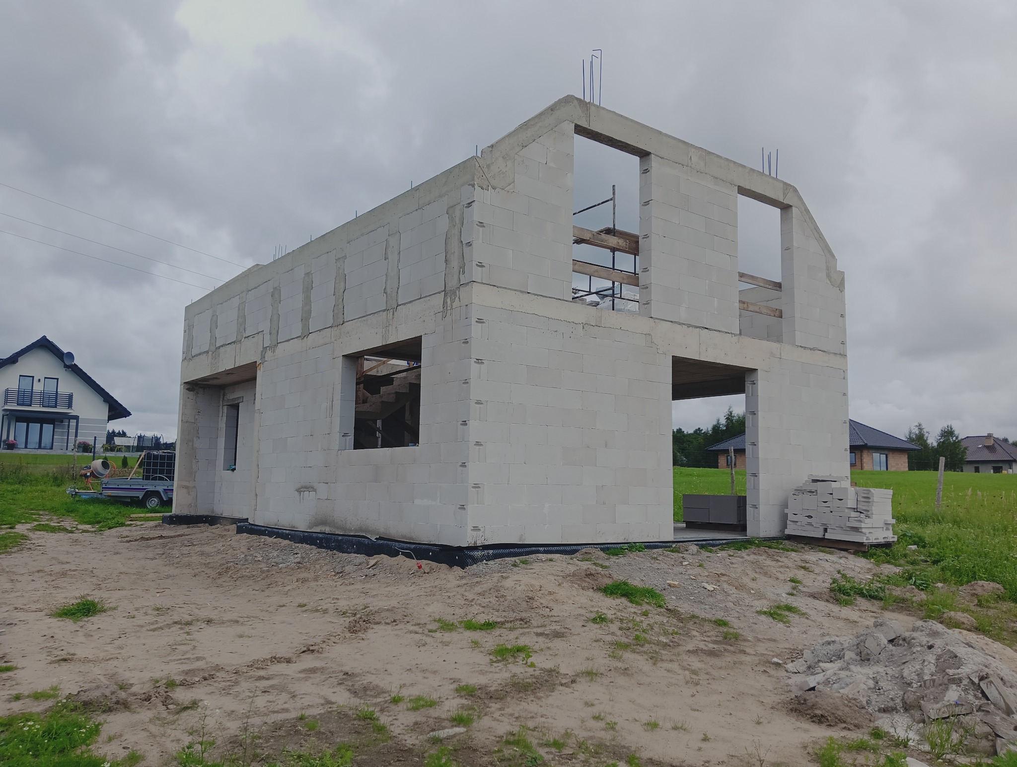 Surowy stan domu z betonu komórkowego w trakcie budowy, widoczne otwory okienne i drzwiowe, zbrojenia, paleta z bloczkami. W tle domy jednorodzinne i pochmurne niebo.