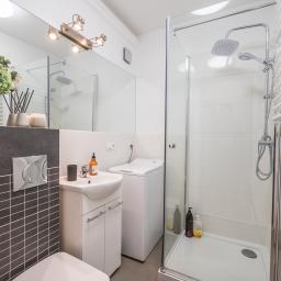 TOMAR BUD Wrocław - Nowoczesna, jasna łazienka z kabiną prysznicową, umywalką z szafką, pralką i dekoracyjnymi płytkami w odcieniach szarości.