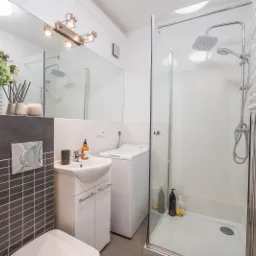 Nowoczesna, jasna łazienka z kabiną prysznicową, umywalką z szafką, pralką i dekoracyjnymi płytkami w odcieniach szarości.