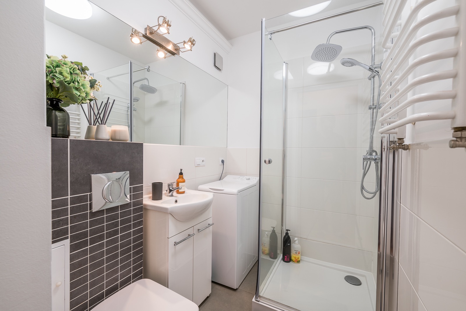 Nowoczesna, jasna łazienka z kabiną prysznicową, umywalką z szafką, pralką i dekoracyjnymi płytkami w odcieniach szarości.
