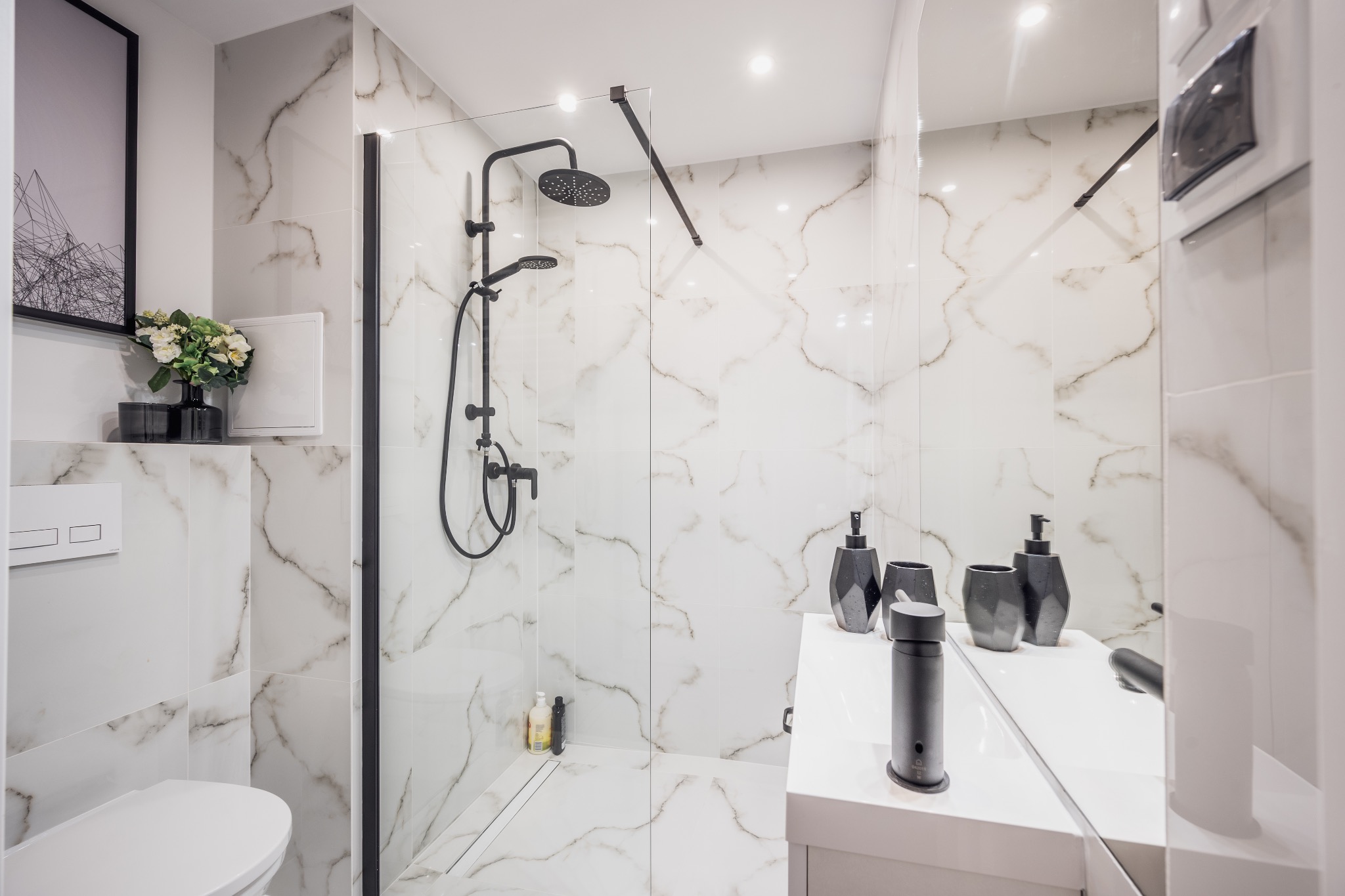 Nowoczesna łazienka z białymi płytkami imitującymi marmur, czarną armaturą, szklanym panelem prysznicowym i minimalistycznymi akcesoriami na blacie umywalki.