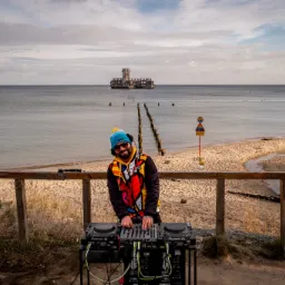 Uśmiechnięty DJ w kolorowej kurtce i czapce, miksujący muzykę na plaży z widokiem na morze i pozostałości molo, w tle żółty znak ostrzegawczy.