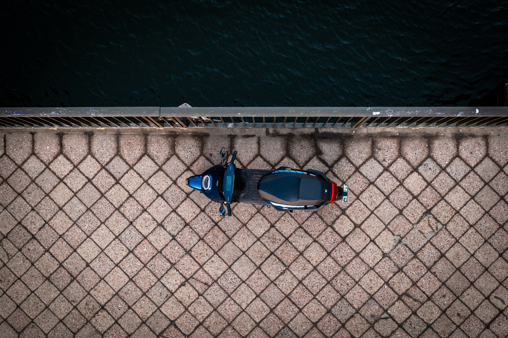 Niebieski skuter elektryczny marki Niu zaparkowany na moście z kostki brukowej, widok z góry, obok ciemna woda.