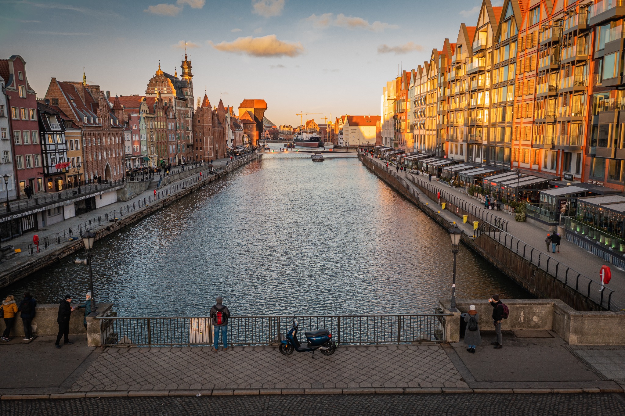 Panorama Gdańska z Motławą w złotym świetle zachodu, widok z mostu z ludźmi i skuterem, charakterystyczna architektura i nabrzeże.