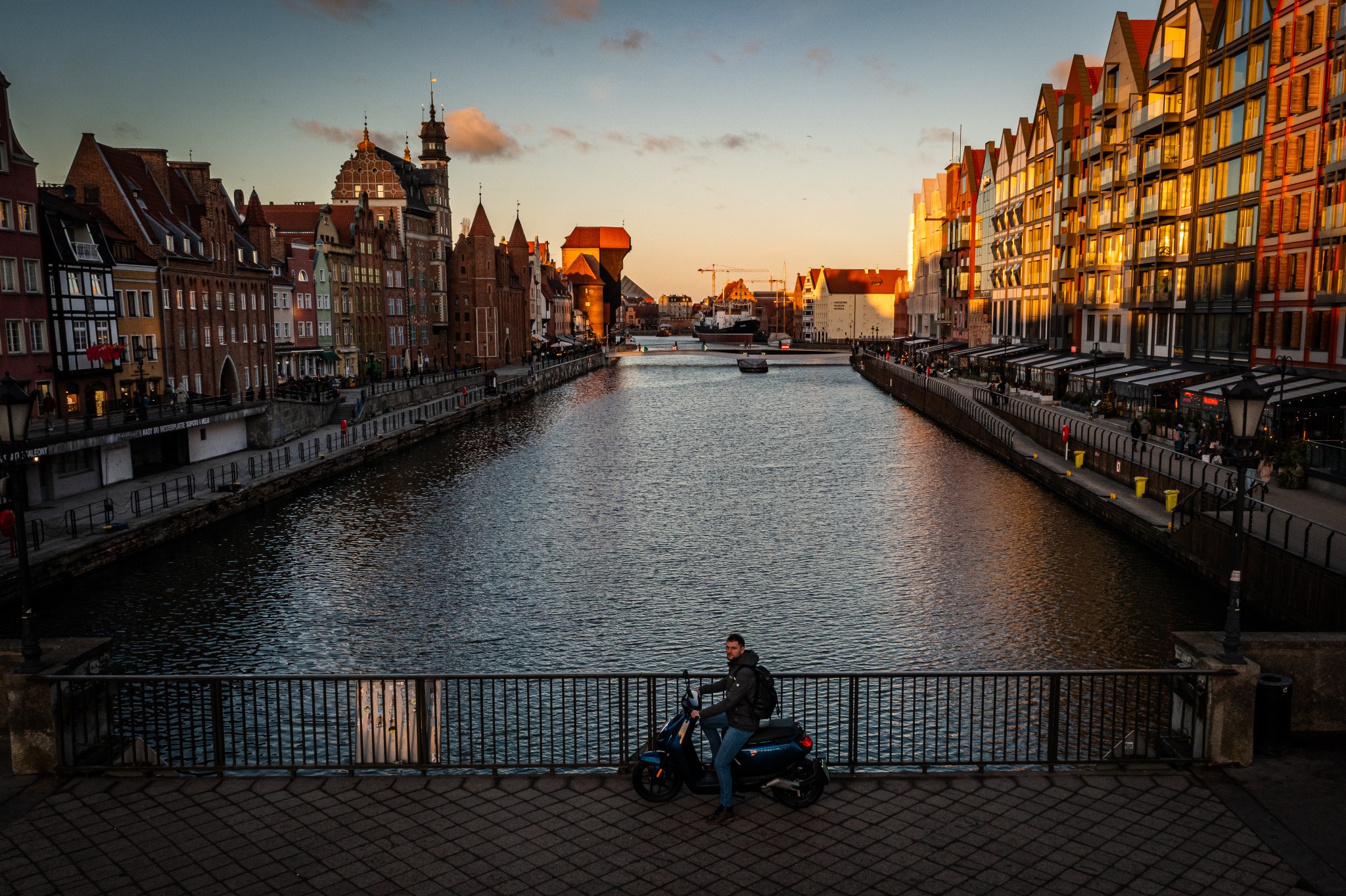 Panorama Gdańska o zachodzie słońca, widok na Motławę z nabrzeżami i zabytkowymi kamienicami, mężczyzna na skuterze elektrycznym na pierwszym planie.
