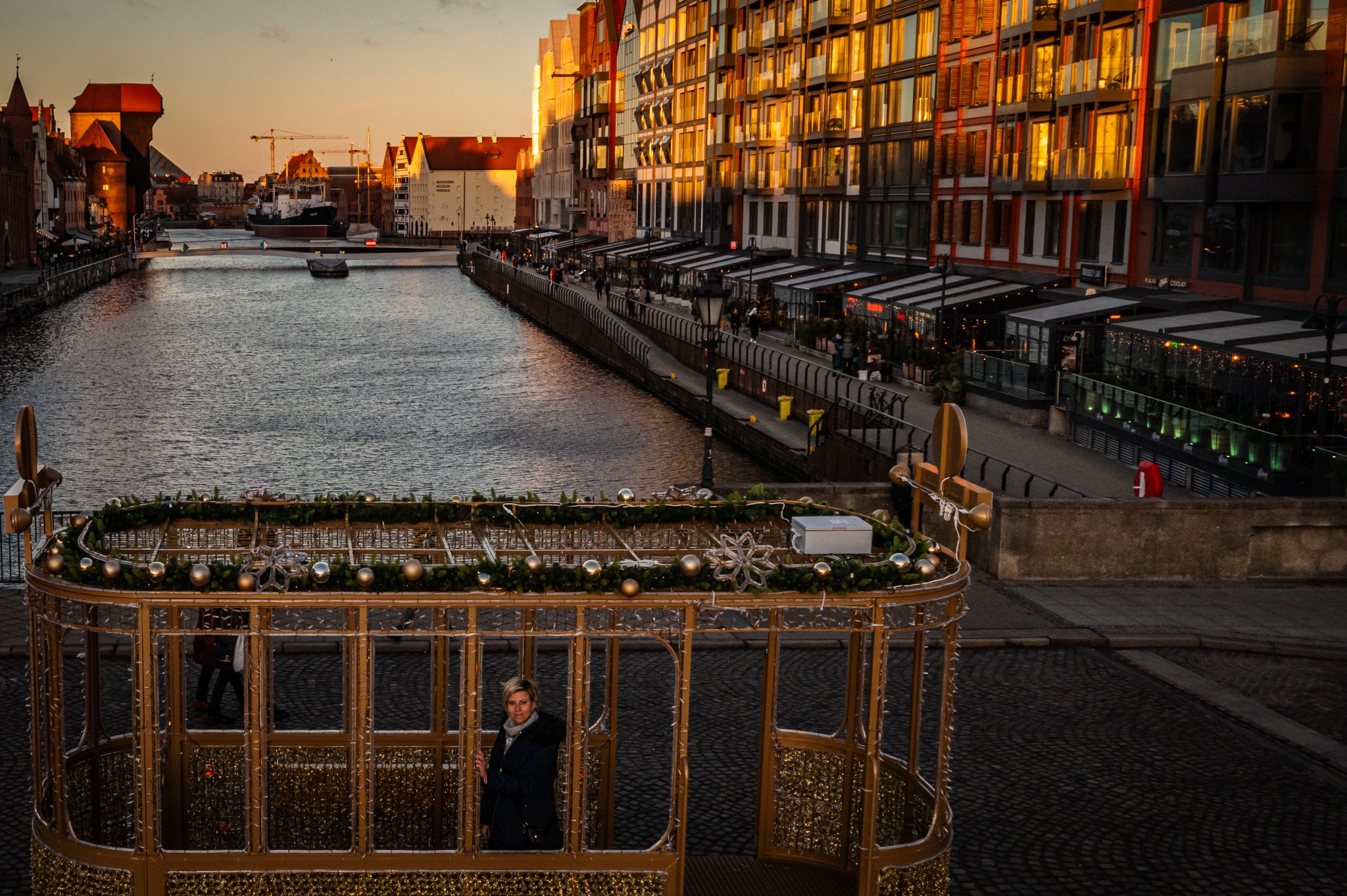 Panorama Gdańska o zachodzie słońca, widok z dekoracyjnej, złotej konstrukcji przypominającej wagonik, w której stoi kobieta w ciemnym płaszczu.