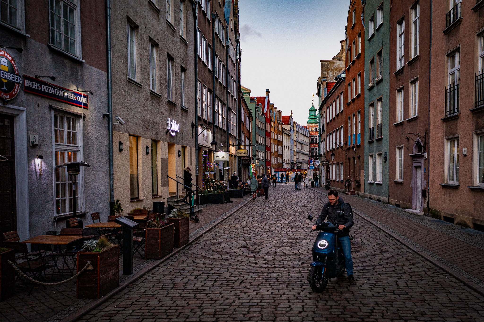 Ulica w Gdańsku z brukowaną nawierzchnią, w oddali widoczna wieża kościoła, a na pierwszym planie mężczyzna na skuterze elektrycznym.