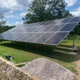 Instalacja paneli słonecznych na konstrukcji wsporczej na trawniku, widoczny mur z kamienia i stos żwiru w tle drzewa i błękitne niebo z chmurami.