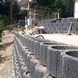 Wznoszenie muru oporowego z szarych, betonowych bloczków z półokrągłymi wycięciami, widoczna taczka, węże i narzędzia budowlane na tle domu i zieleni.