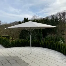 Duży, kwadratowy parasol ogrodowy na patio z jasnoszarej kostki, otoczony żywopłotem z tuj.